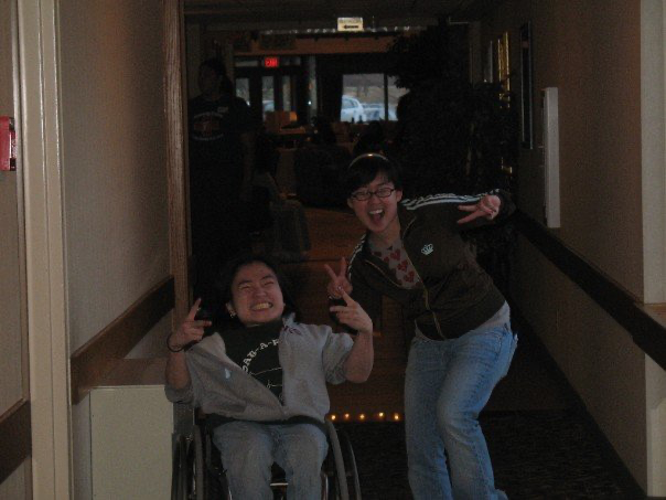Photo of Mia at around age 20. She has long hair that is in a ponytail and she uses a wheelchair. Next to her is her friend Catherine, who is also Asian American. She has short black hair and glasses. Both pose for the camera gesturing with peace signs on both hands.