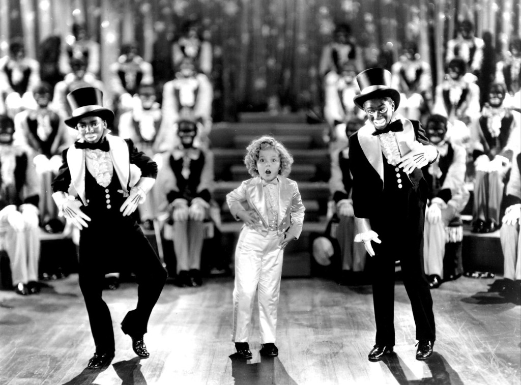 Shirley Temple dances with two men in Blackface, while other actors also in Blackface look on, from Dimples (1936).