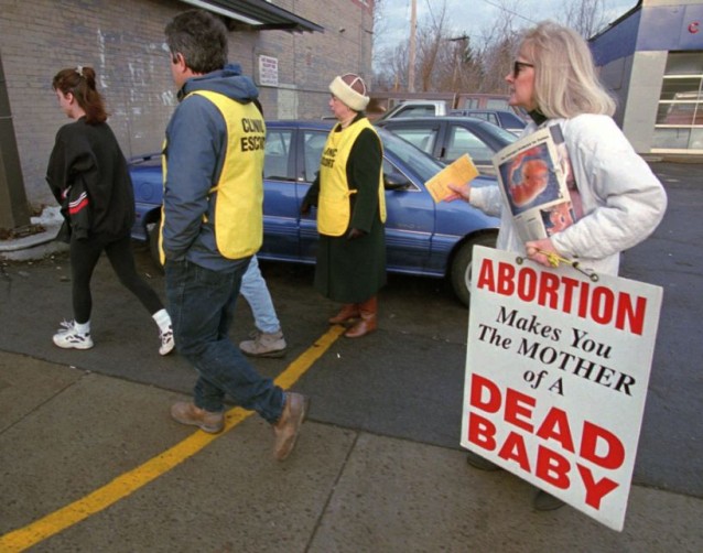 An anti-choice protester harasses a woman and her clinic escorts as she crosses the yellow line into the clinic buffer zone. Photo credit: ThinkProgress