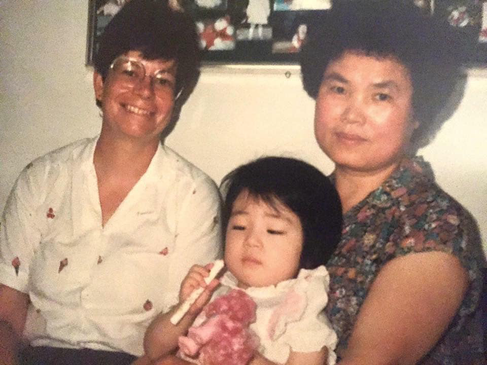 Photo of Mia at 3-years-old, with short black hair and bangs. She sits on her foster mother's lap, looking at the end of a pen light. Her foster mother is Korean. She has a pink My Little Pony in her other hand. Sitting next to her foster mother is her adoptive mother, who is white.